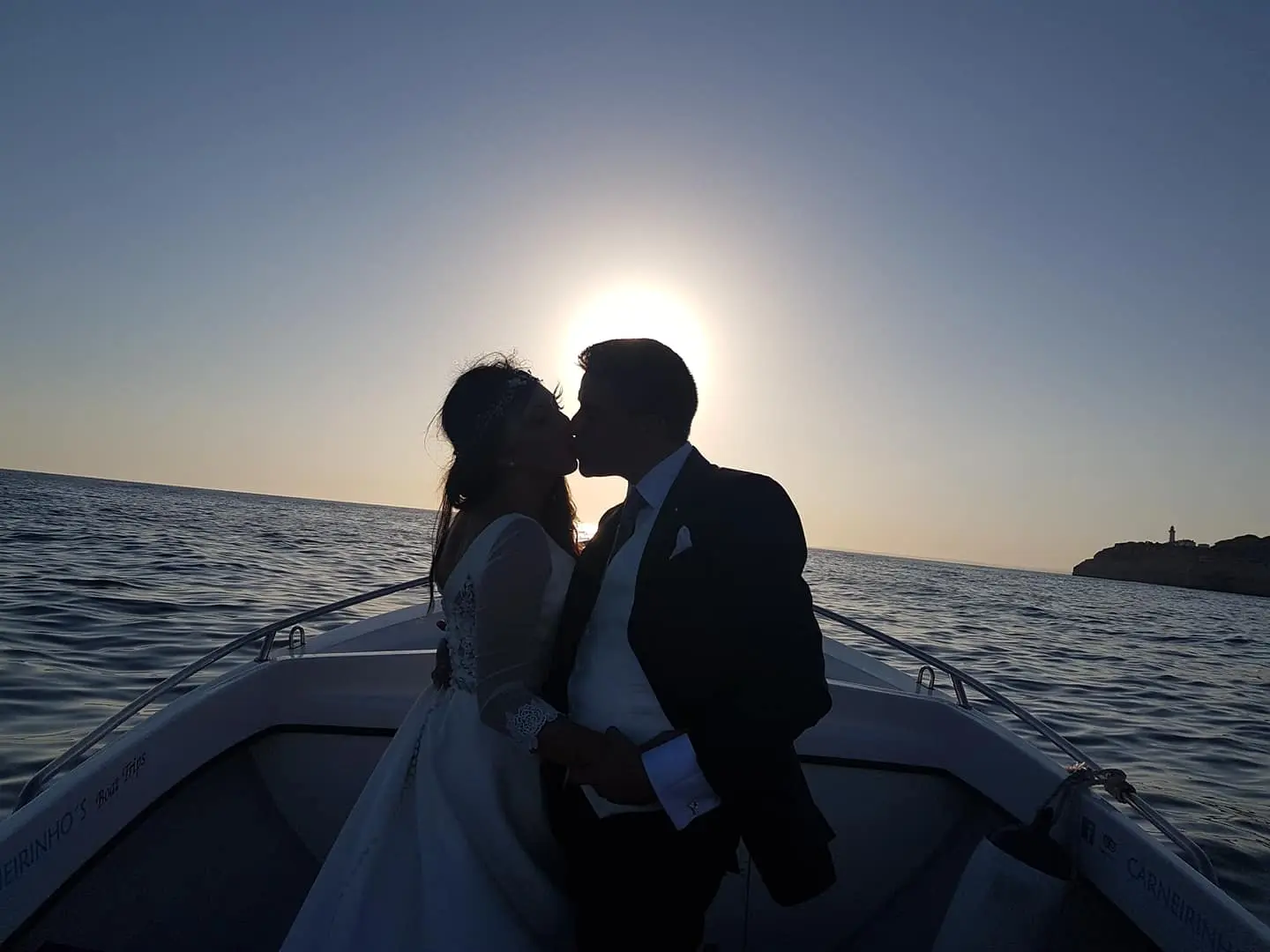 Casal em silhueta na proa ao pôr do sol, com um farol à direita